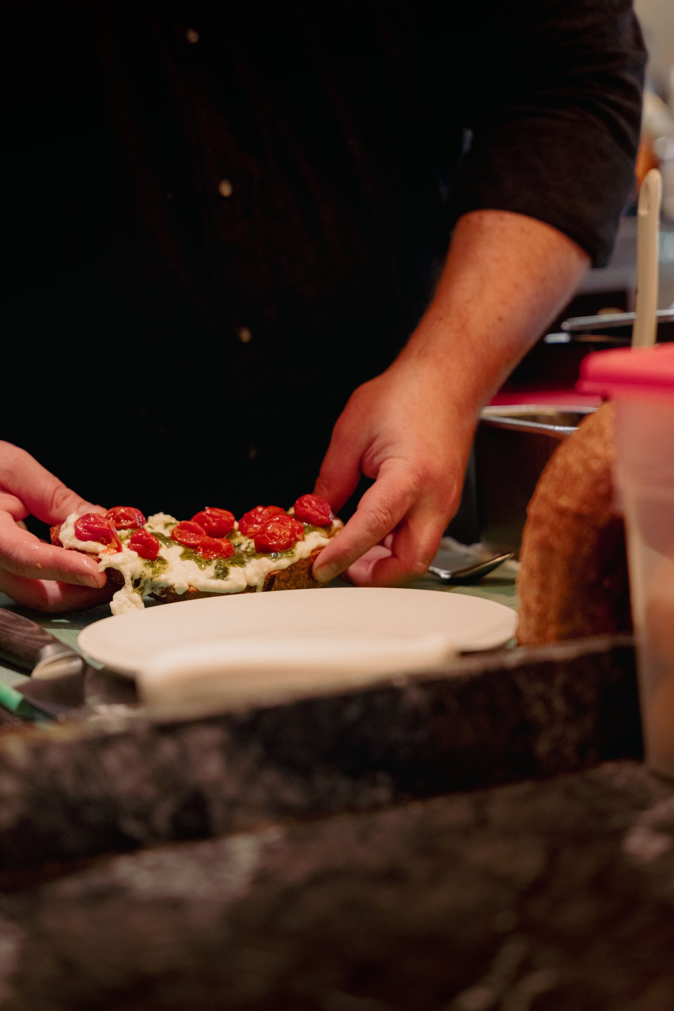 Chef hands plating artisanal toast topped with creamy burrata, herb pesto, and blistered roasted cherry tomatoes under focused warm kitchen light